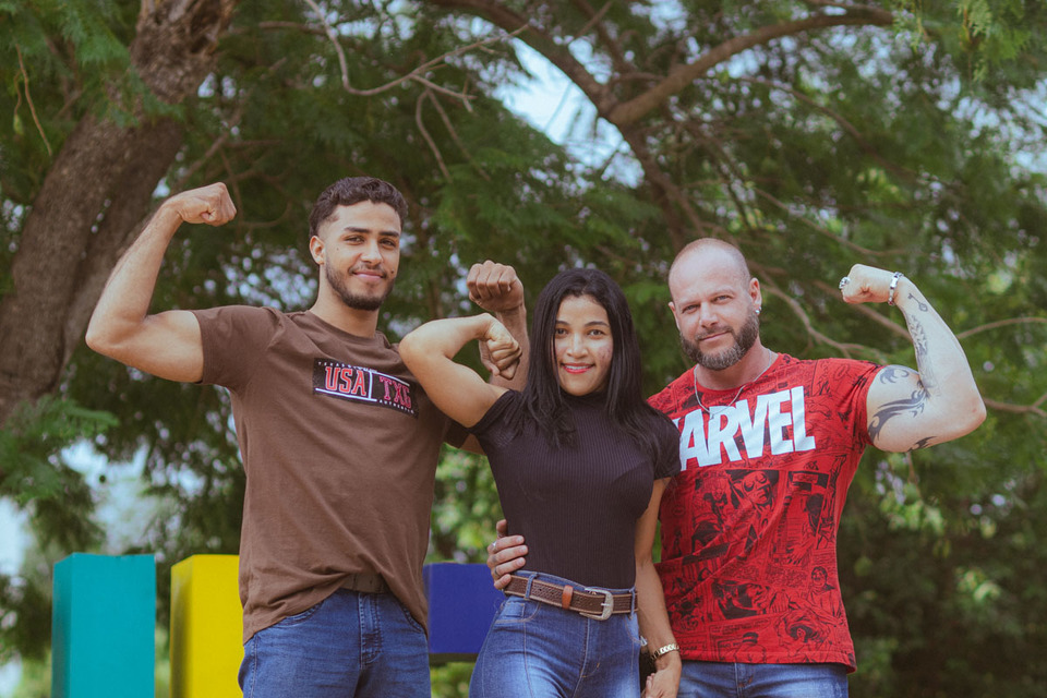 Dois homens fortes fazem pose para mostrar os biceps ao lado da estudante que repete a pose